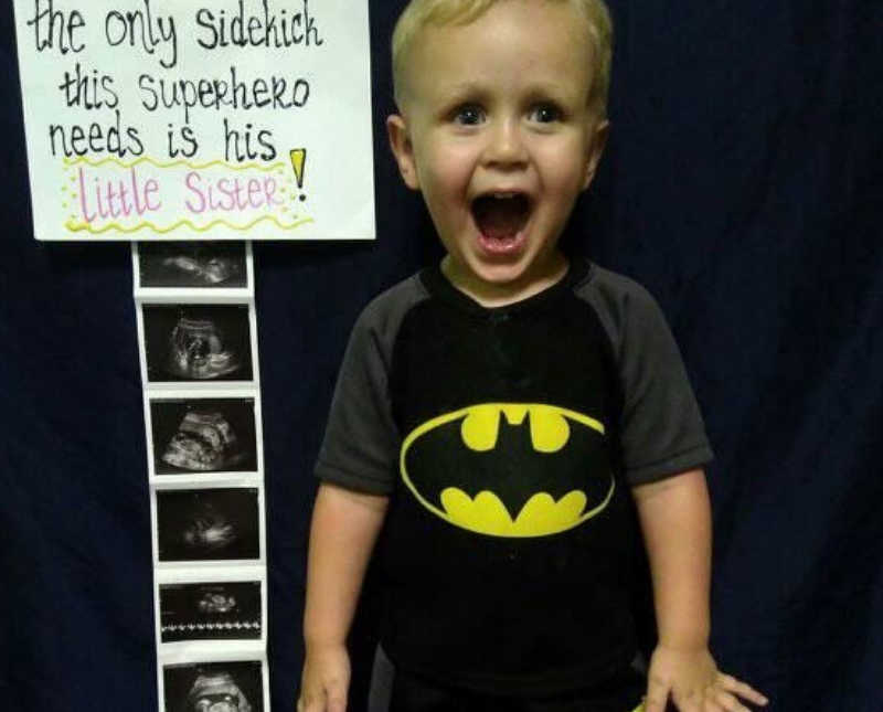 Little boy stands smiling with mouth open beside sign announcing baby sister and ultrasound pictures of her