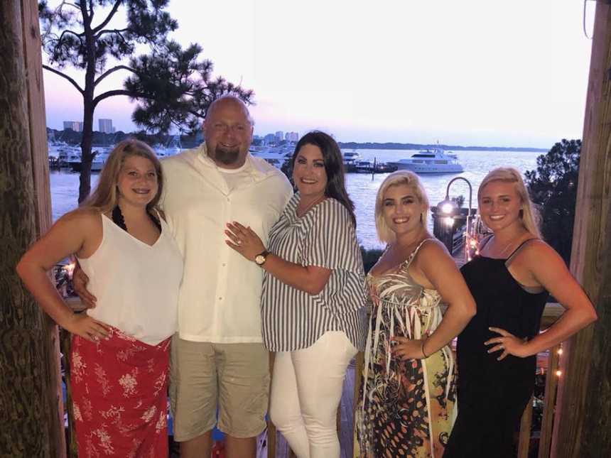 Husband and wife stand with their three girls with body of water in background