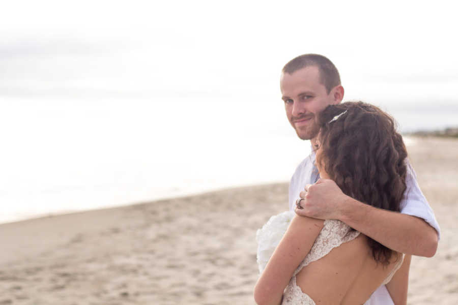 Groom smiles as he rests arm around bride as they stand on beach