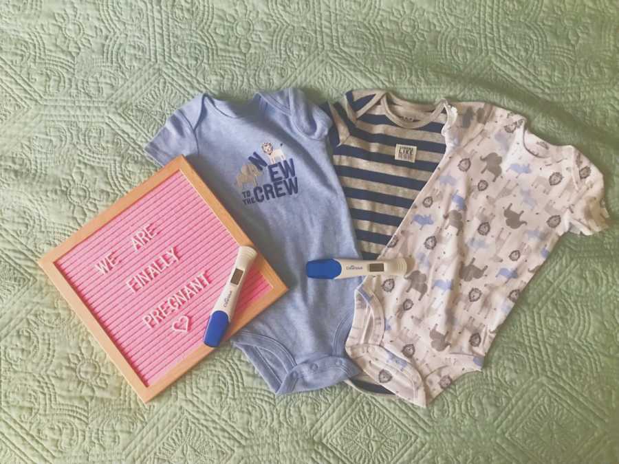 Three onesies lay on green blanket beside sign announcing pregnancy and two pregnancy tests