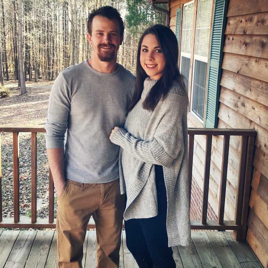 Husband and wife stand smiling on porch of wood house