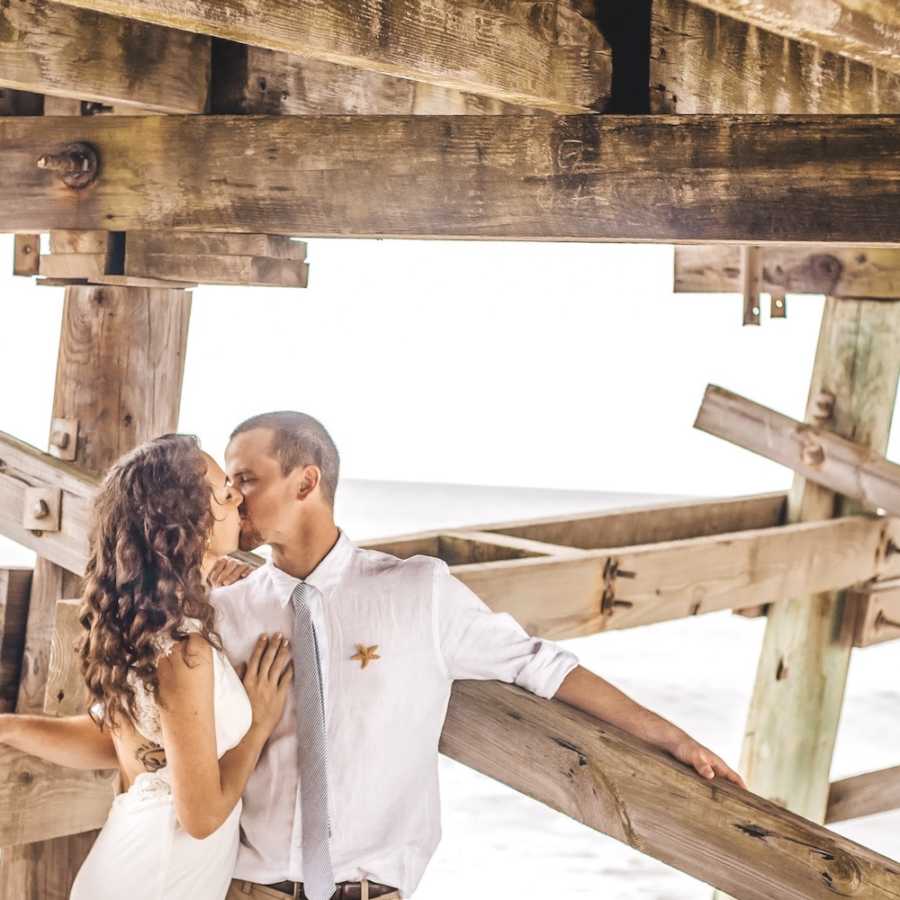 Bride and groom stand kissing under pier