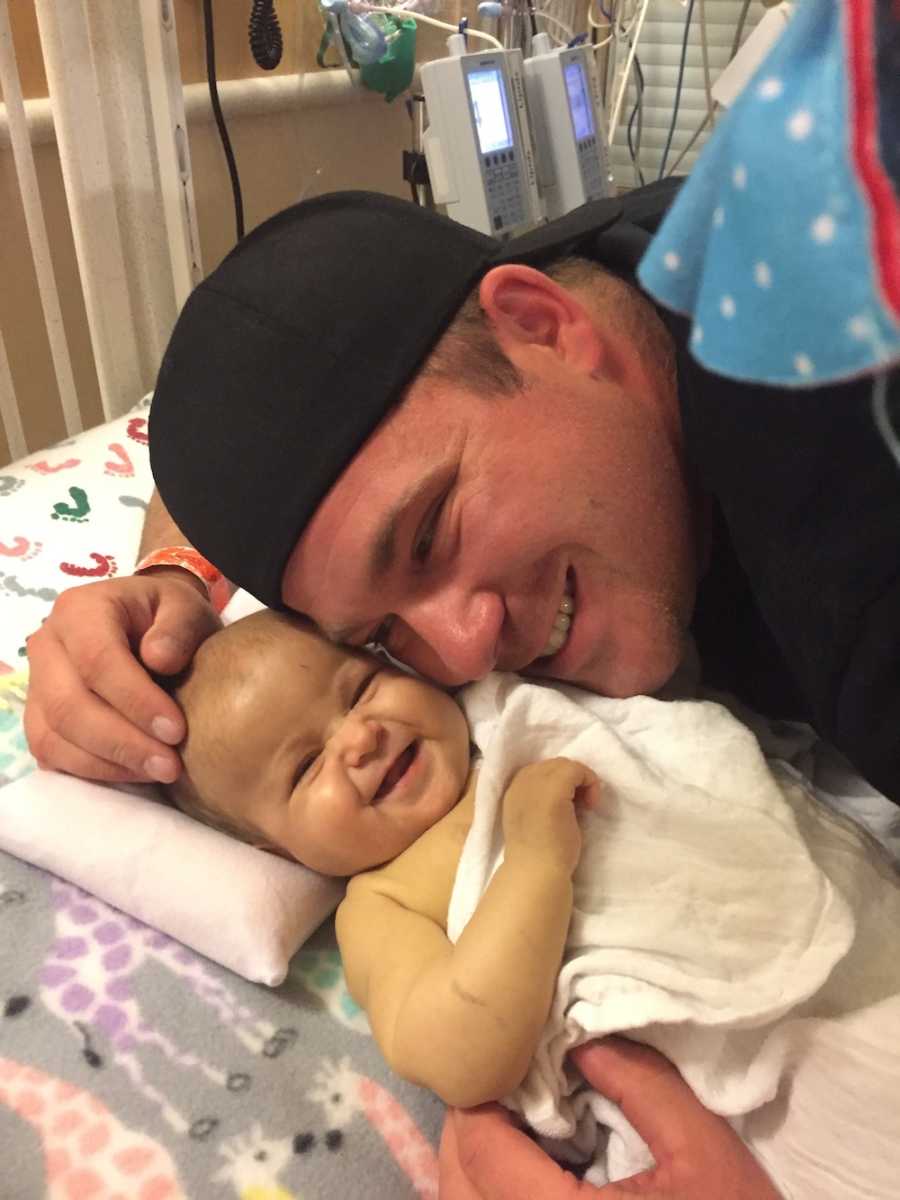 Father leans over to hug smiling baby in hospital bed who has heart defect