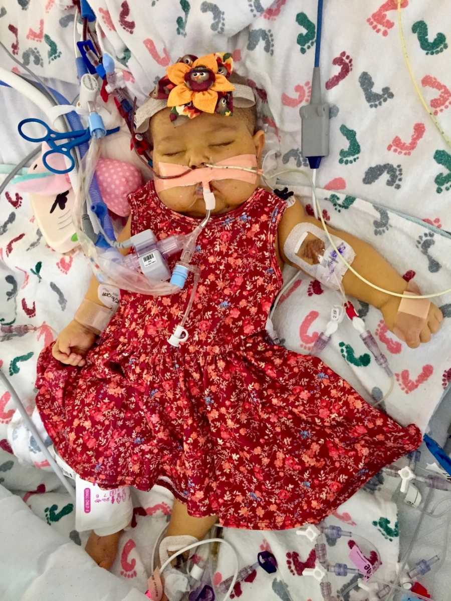 Baby who is awaiting transplant lays in hospital bed in red dress with tubes and wires attached to her