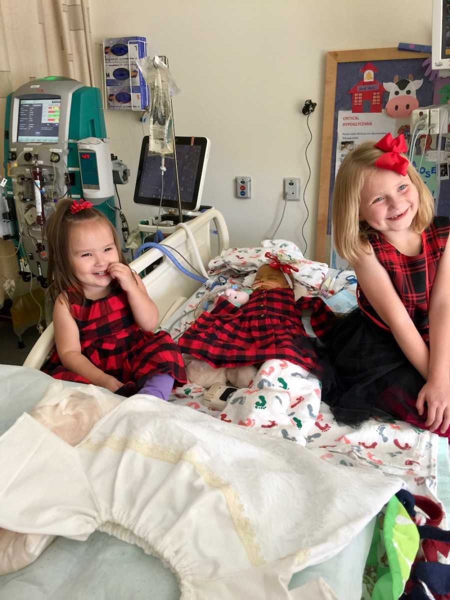 Older sisters sit in hospital bed where baby sister who needs heart transplant lays
