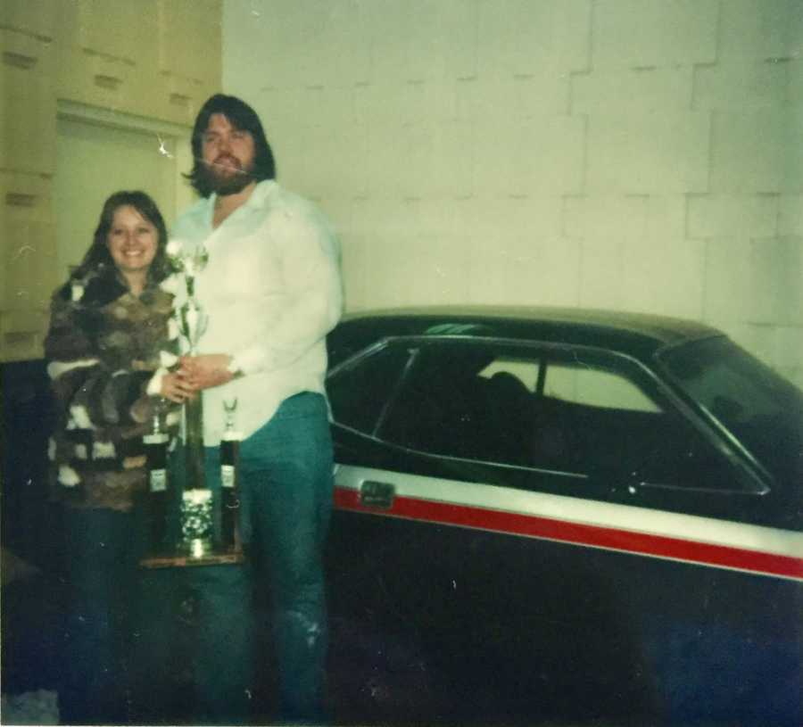 Late husband and wife stand beside car holding trophy