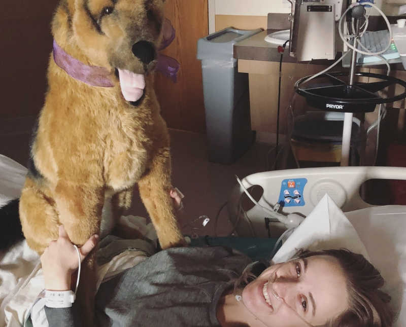 Young woman who fell off horse lays smiling in hospital bed with large stuffed dog sitting on her chest