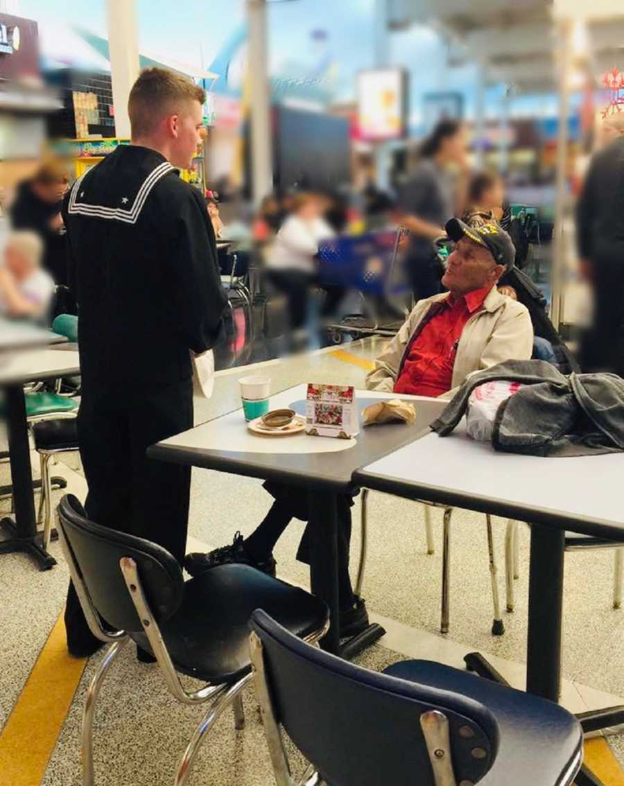 Navy seal stands beside table at food court where veteran sits