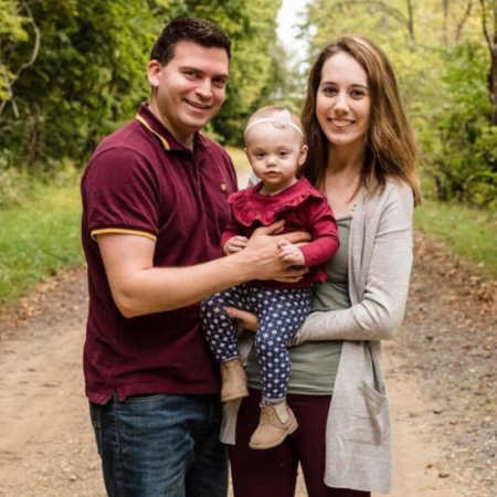 Husband and wife stand in dirt path in wooded area holding their young daughter