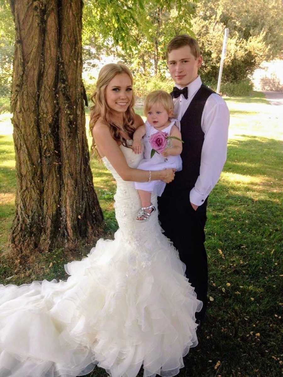 Teen bride and groom stand outside beside tree holding their daughter