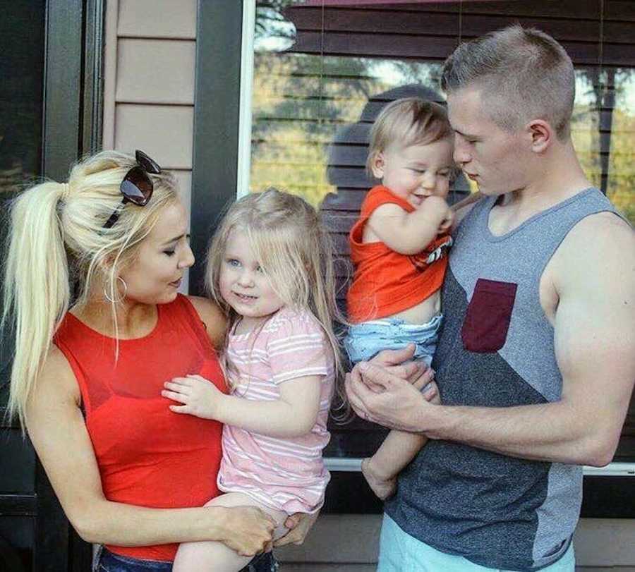 Young husband and wife stand outside home holding son and daughter