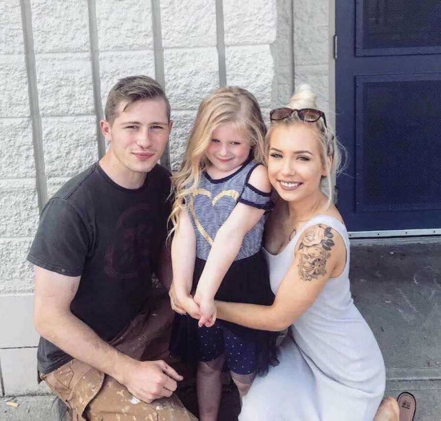 Young husband and wife kneel on either side of their daughter who stands in between them