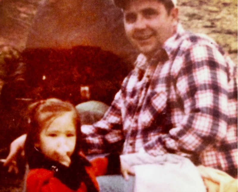 Little girl stands sucking her thumb beside father who sits smiling