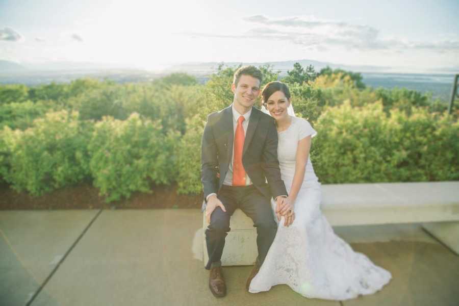 Bride and groom sit on bench outside with greenery in background