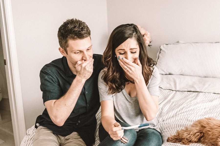 Husband and wife sit on bed with hands over their mouth as they look at positive pregnancy test
