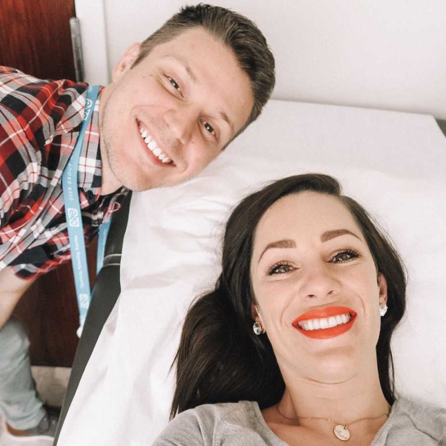 Woman smiles in selfie at fertility clinic with husband beside her