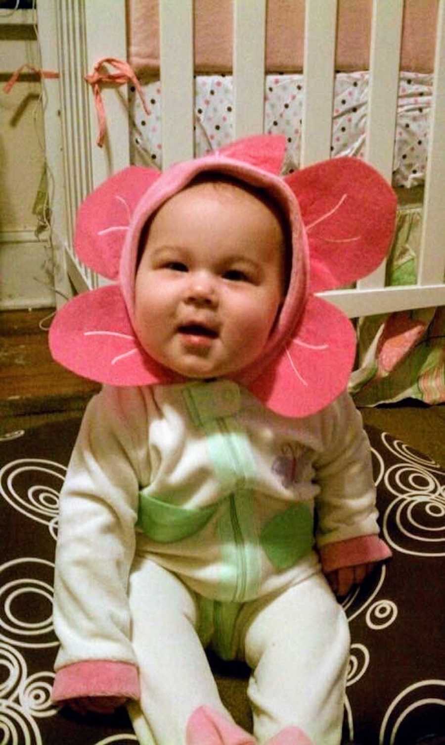 Baby girl sits on floor wearing onesie that has flower petals that goes around her head