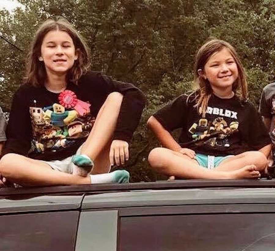 Two young girls sit smiling on roof of car