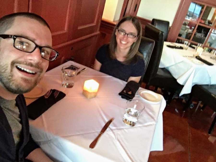 Man smiles as in selfie with wife who sits across the table from him at restaurant 