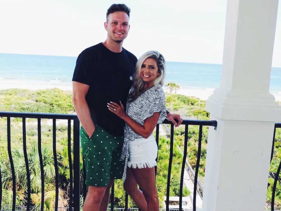 Husband and wife stand smiling beside railing that looks out to beach