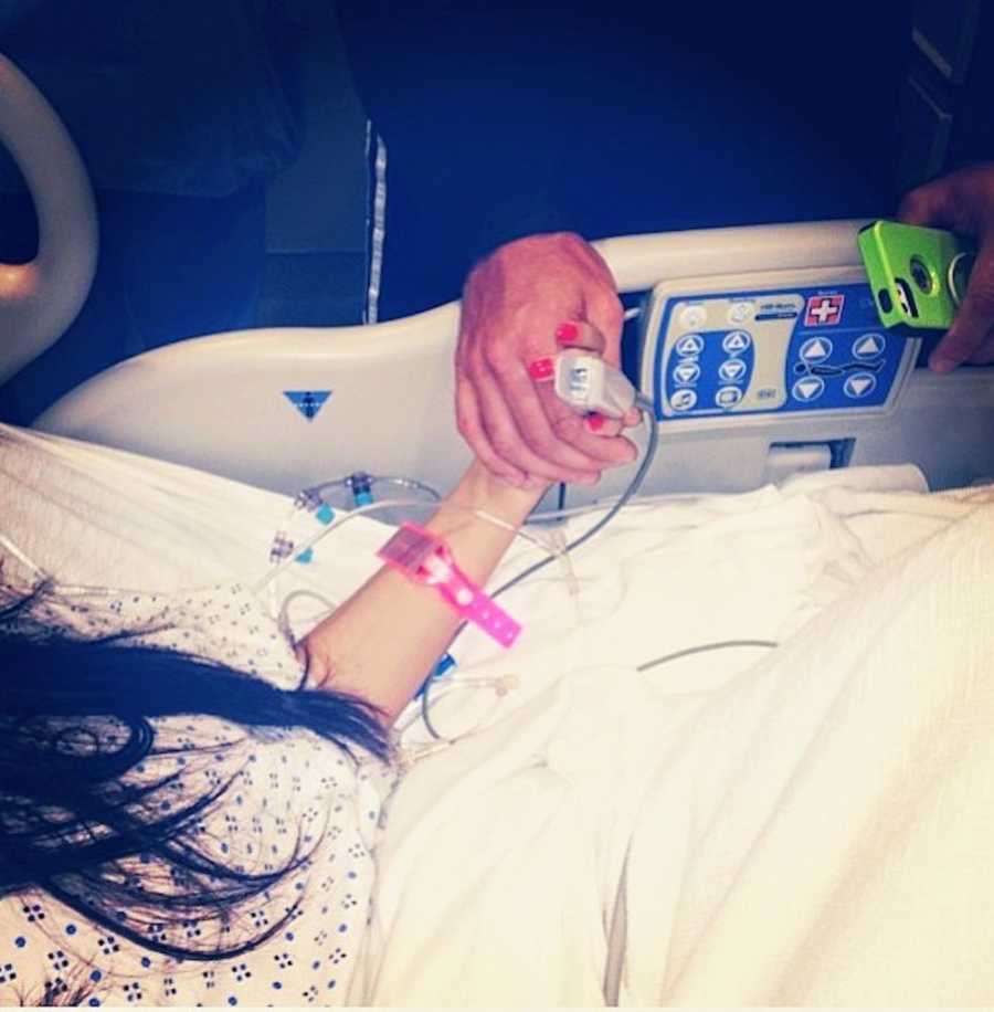 Close up of woman laying in hospital holding hands with someone who stands over her