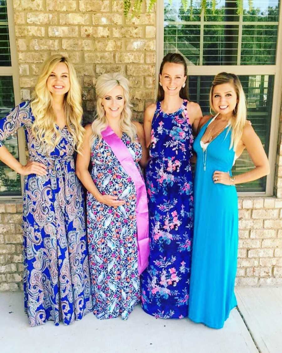 Pregnant woman stands holding her stomach beside three woman in long dresses