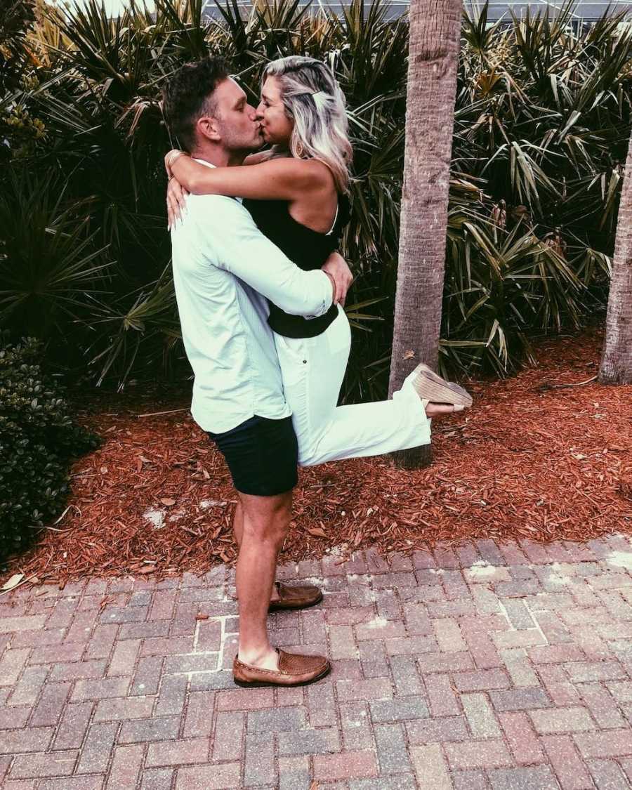 Husband holds wife up as he kisses her beside tropical plants