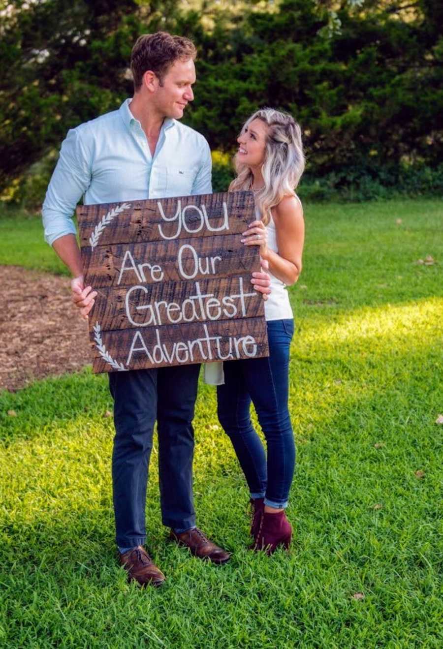 Husband and wife stand outside holding sign that says, "you are our greatest adventure"