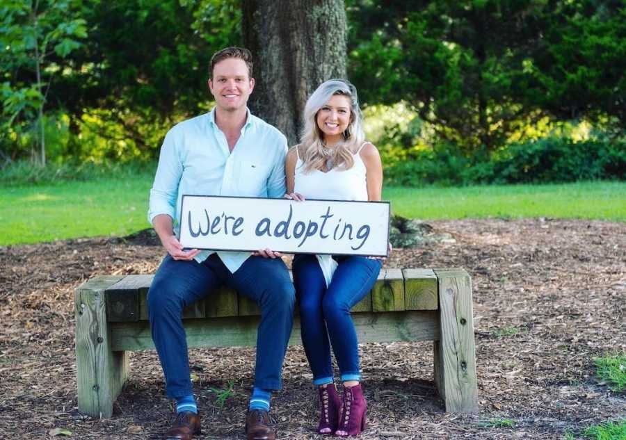 Husband and wife sit on bench outside holding sign that says, "we're adopting"