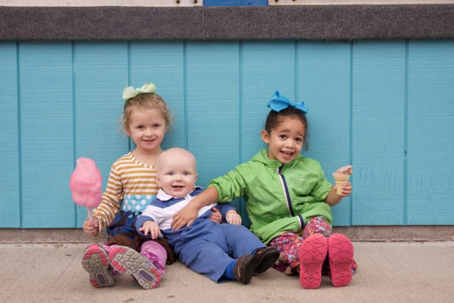 Little girls sit on ground with ice cream and cotton candy with their baby brother between them