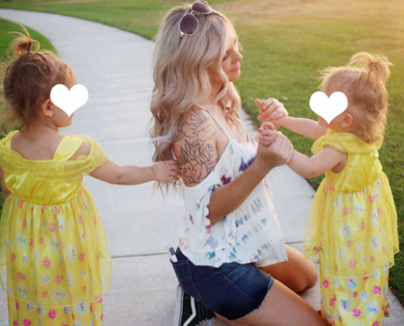 Foster mother kneels on sidewalk holding hands of little girl while another little girl stands behind her