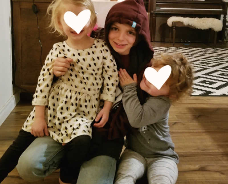 Foster child sits on floor of home with two young foster sisters beside him