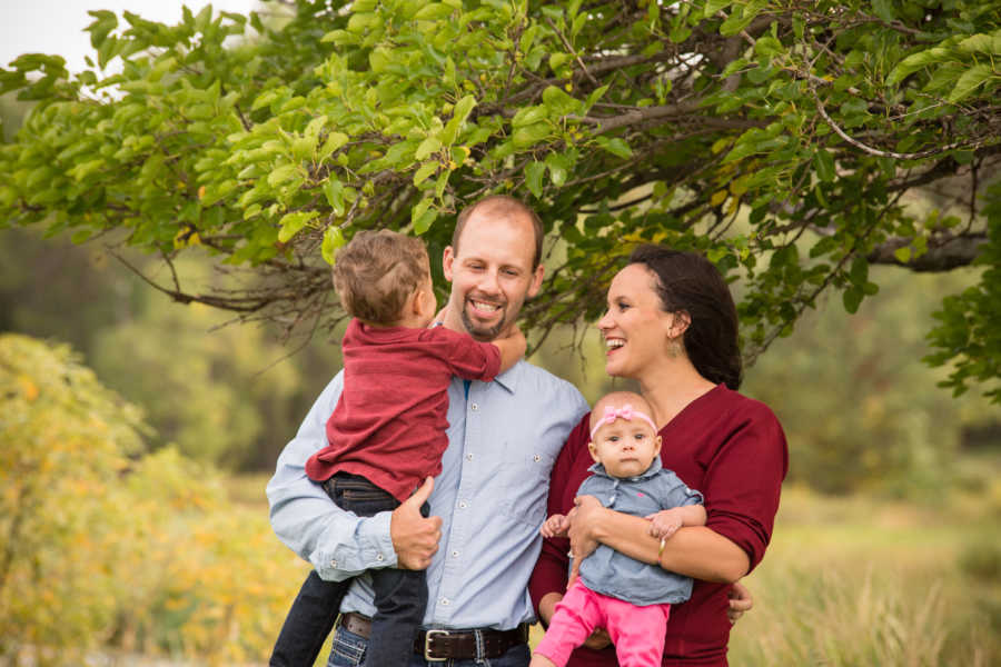 Husband stands outside holding son beside wife who holds their baby daughter