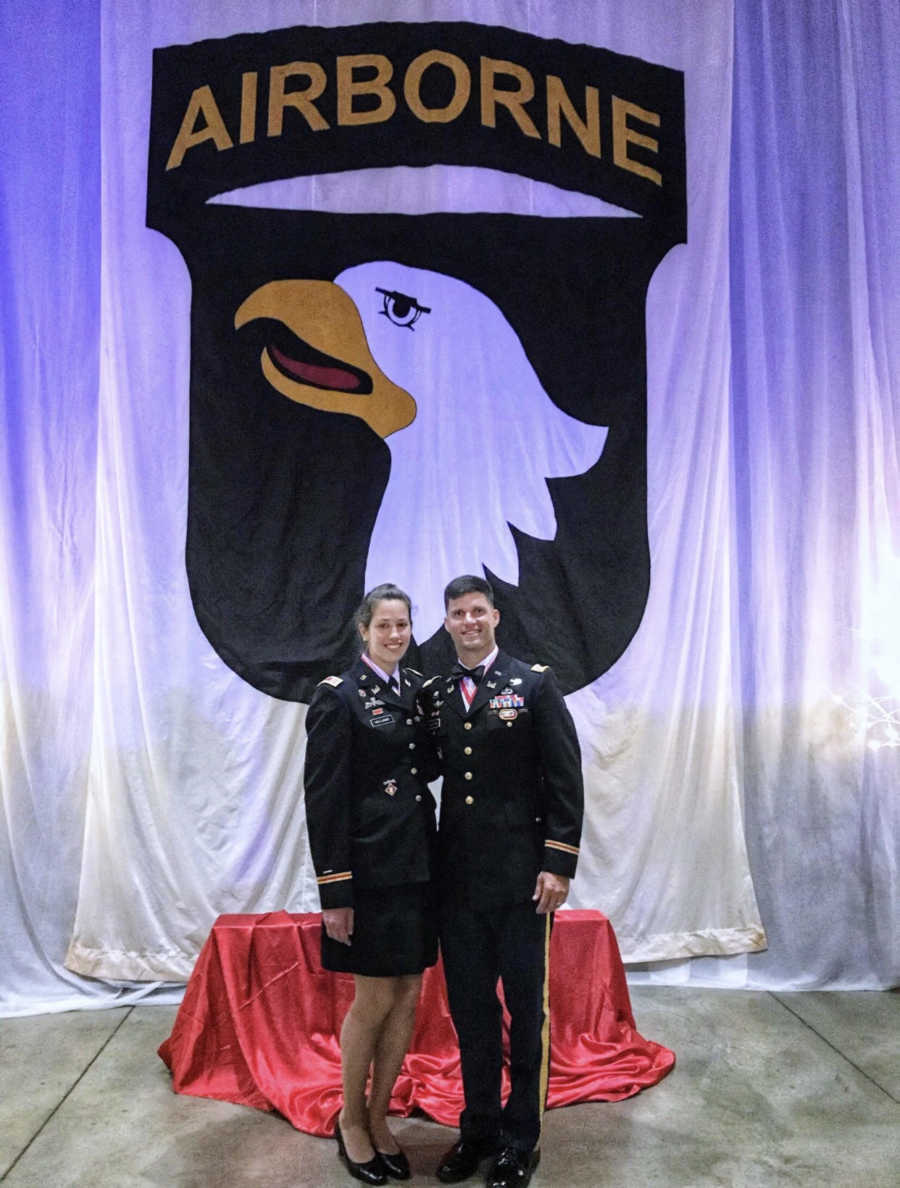 Husband and wife who are active duty in army stand in uniform in front of backdrop that says, "Airborne"