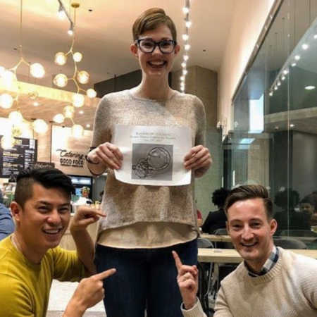 Surrogate stands smiling holding picture of implanted embryo while gay couple smiles pointing at her stomach