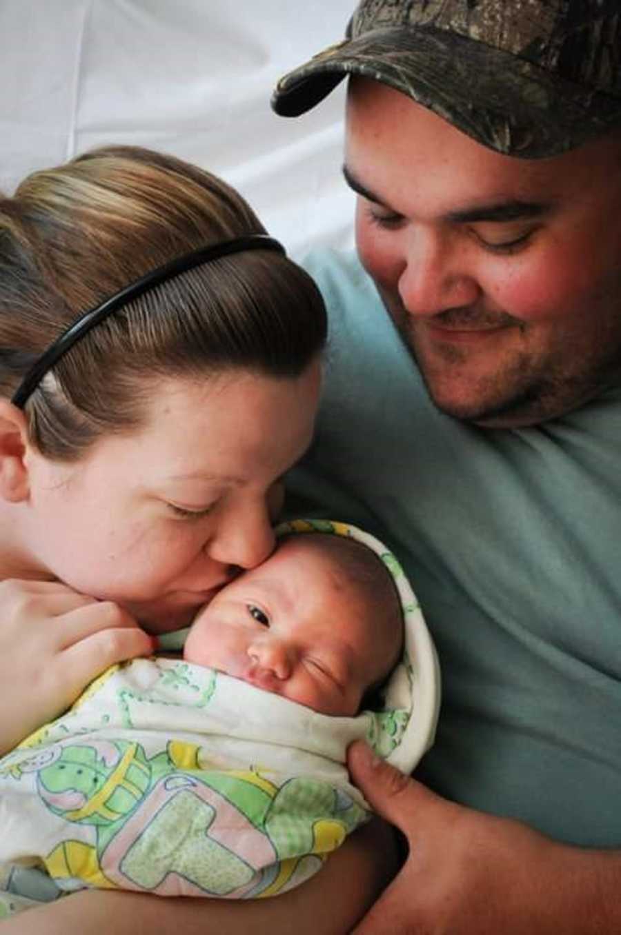 Mother kisses newborn baby's head that is held in her arms while husband sits next to her