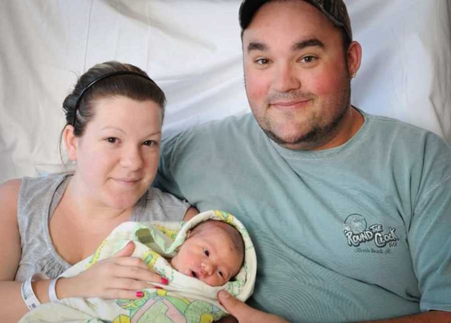 Husband and wife smiles as they sit holding newborn wrapped in blanket
