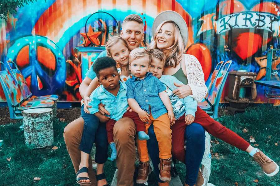 Husband and wife sit in chair outside with their three biological kids and adopted son in their laps