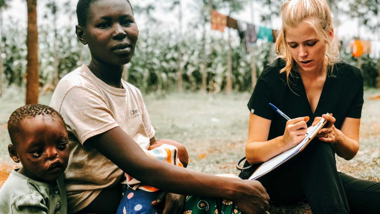 Woman sits on ground of orphanage writing notes beside mother and daughter with facial disfiguration