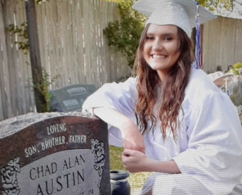 Young woman in white cap and gown kneels beside man's head stone