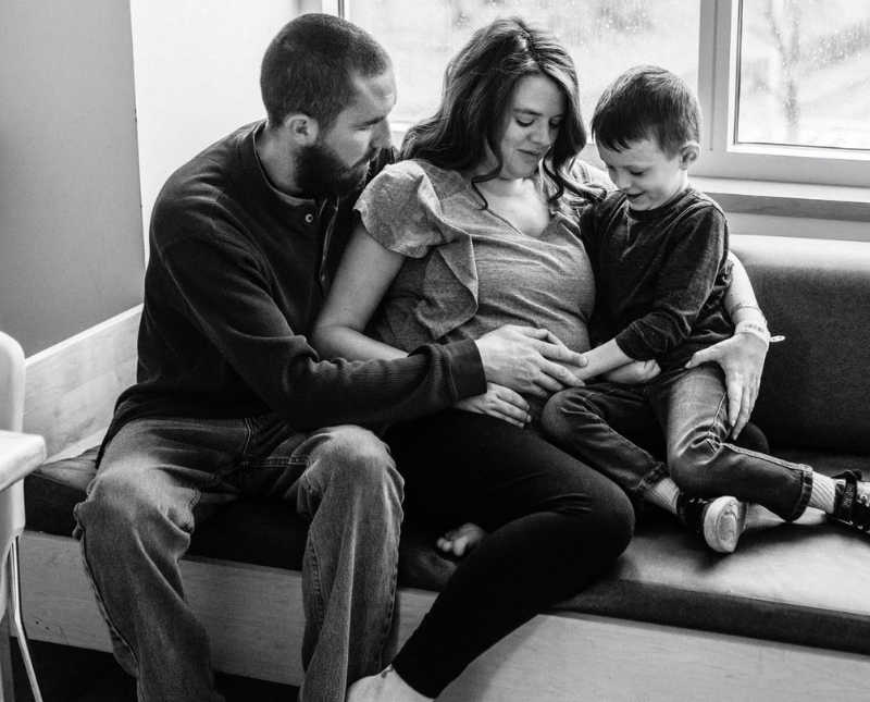 Pregnant woman sits on couch in hospital room while her son and husband touch her stomach