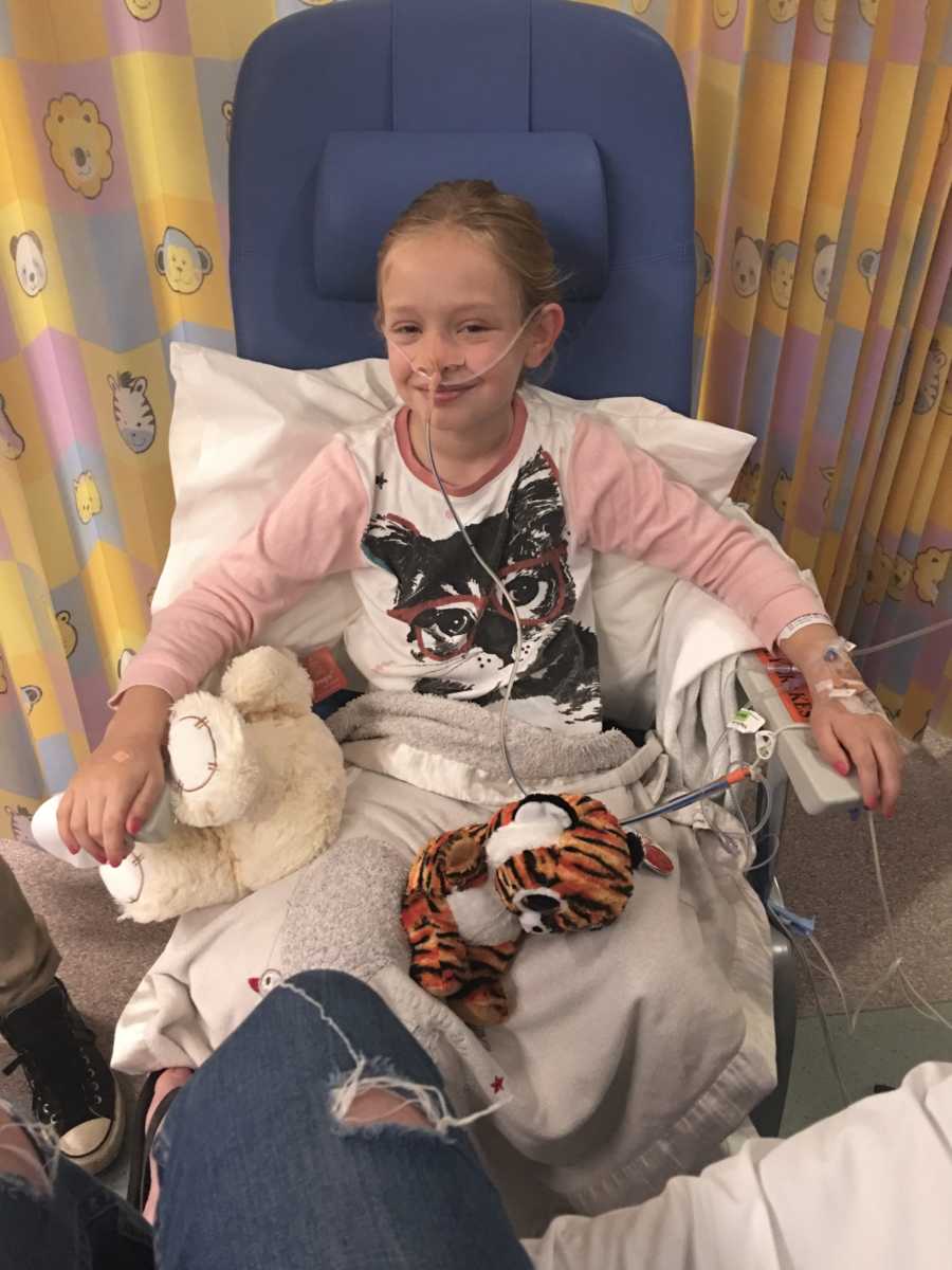 Little girl sits in hospital room with tube up her nose and stuffed animals on her lap
