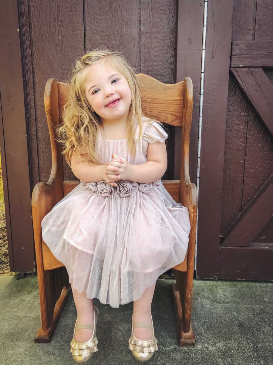 Little girl with down syndrome sits smiling in chair wearing beige dress