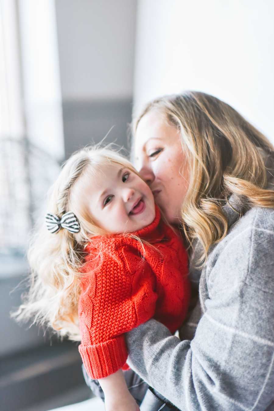 Little girl with down syndrome smiles as her mother holds her and kisses her on cheek