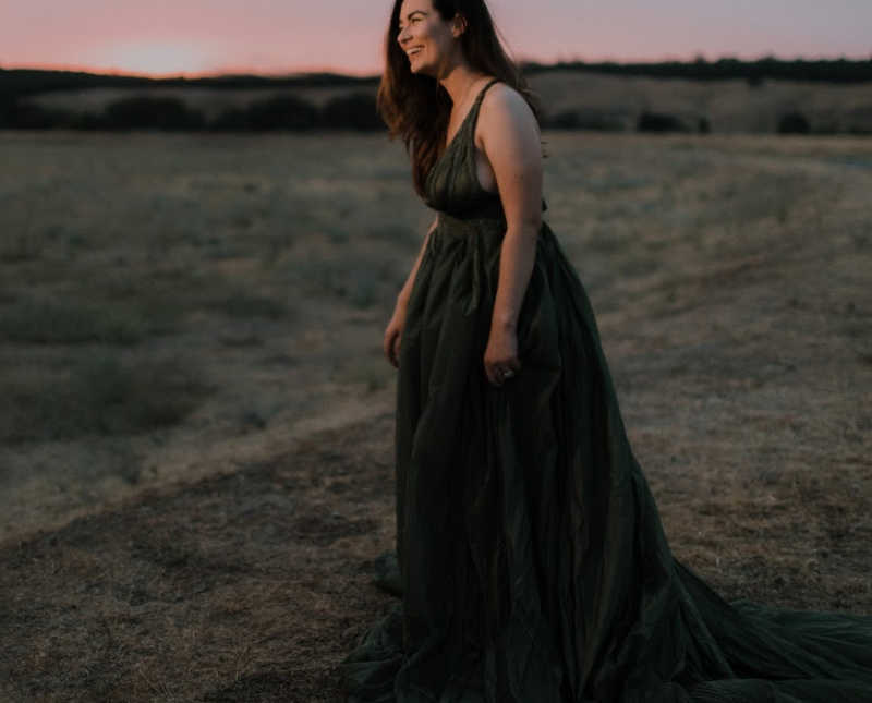 Woman who had hysterectomy stands outside at sunset smiling in long dress