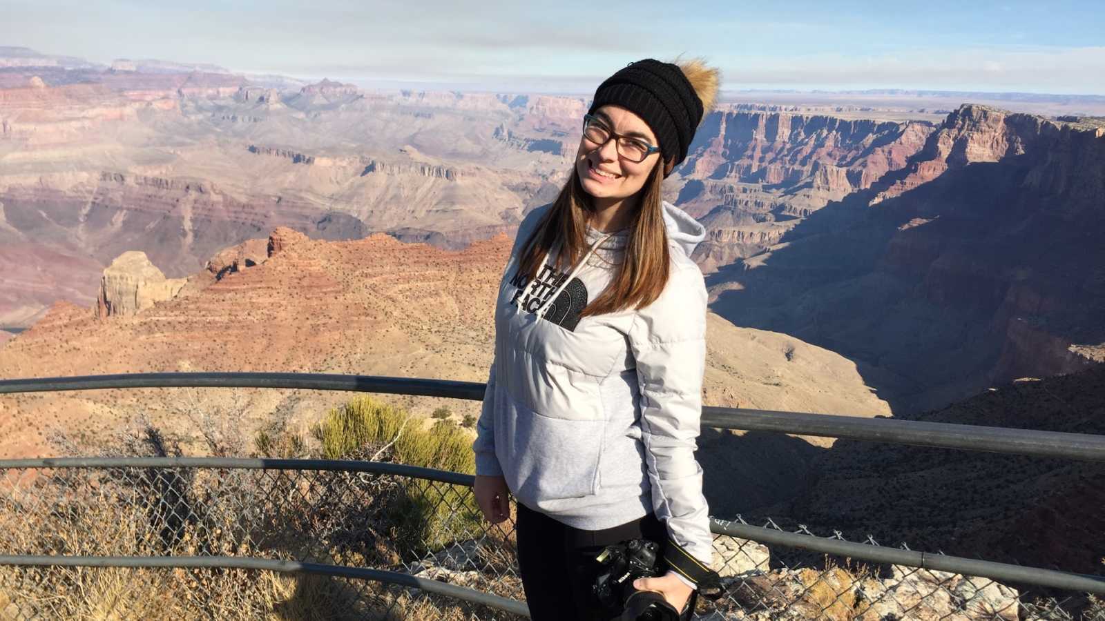 Young woman stands smiling beside railing that over looks Grand Canyon