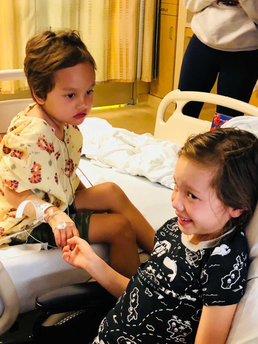 Young brother and sister with brain cancer sit in hospital room holding hands