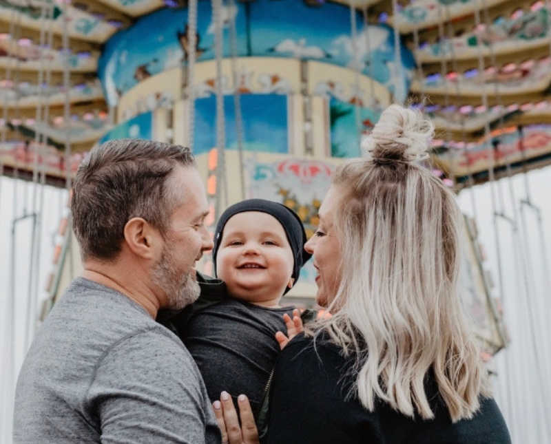 Husband and wife stand holding baby son with carnival ride in background