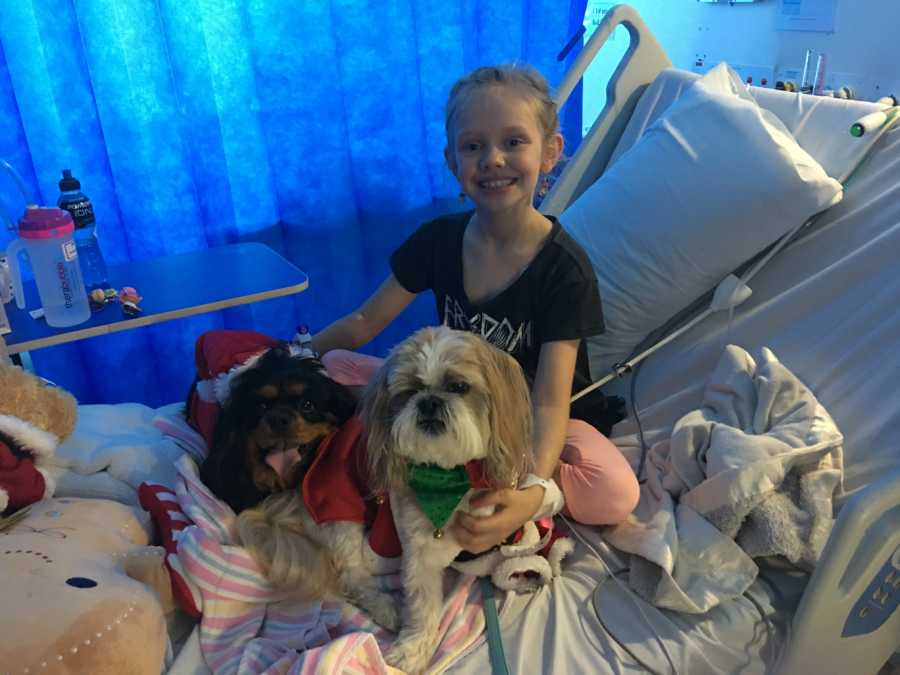 Little girl with rhinovirus sits smiling in hospital bed with two little dogs in bed with her