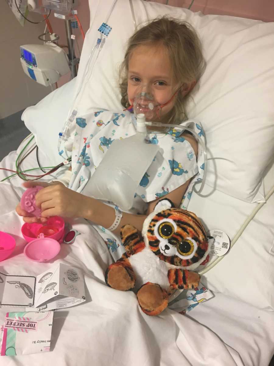 Little girl with lung issues lays in hospital bed smiling as she plays with toys